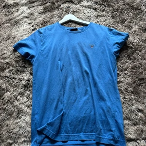 Blå t-shirt från GANT i bomull - Blå t-shirt från GANT med klassisk rund hals och korta ärmar. Liten broderad logga på bröstet ger en clean och stilren look. Tillverkad i mjuk bomull som känns skön mot huden. Perfekt för dig som gillar enkel och snygg design.