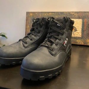Svarta boots från Fila  - Säljer ett par svarta boots från Fila i storlek 39. Skorna har en kombination av skinn och syntetmaterial, med Fila-logga på sidan och på sulan. Perfekta för dig som vill ha en cool och stabil look till hösten och vintern. Dom är använda minst 2 gånger och har en liten repa på skinnet på framsidan men inget som man märker.