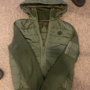 Grön cardigan med huva - Snygg grön pufferjacka med stickade ärmar och ribbade muddar. Jackan har huva, dragkedja framtill och praktiska fickor med dragkedja. Quiltad kropp i syntetmaterial och ärmar i stickad kvalitet. Perfekt för kyliga dagar.