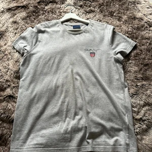 Grå t-shirt från GANT - Snygg grå t-shirt från GANT med klassisk passform och rund hals. Diskret GANT-logga och sköld på bröstet. Tillverkad i mjuk bomull som känns skön mot huden. Perfekt till jeans eller shorts för en clean och enkel stil.