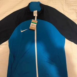 Blå och svart vindjacka från Nike - Sportig vindjacka från Nike i blått med svarta ärmar och vit dragkedja. Jackan har hög krage, lång ärm och klassisk Nike-logga på bröstet. Perfekt för träning eller chill vardagsstil.