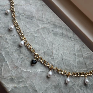 Guldigt halsband med pärlor och hjärta - Unikt halsband med chunky guldfärgad kedja, dekorerat med droppformade vita pärlor och ett svart hjärthänge i mitten. Perfekt statement piece med cool vibe och snygga detaljer som ger extra edge till din look.