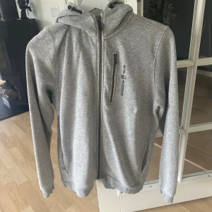 Sailracing zip hoddie  - Snygg grå hoodie från Sail Racing med hel dragkedja framtill och broderad logga på bröstet. Tröjan har huva, lång ärm och en bröstficka med dragkedja. Perfekt för dig som gillar sportig och clean stil. Koftan har en liten sminkfläck på armen, men inget som syns eller stör.