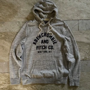 Abercrombie & Fitch hoodie  - Snygg gråmelerad hoodie från Abercrombie & Fitch med mörkblå tryck på bröstet. Tröjan har huva med vita snören. Perfekt för chill dagar och har en normal fit som sitter skönt. Obs något missfärgat vid kragen och 2 svaga fläckar på magen, svårt att se på bild!Modell 188cm! Mvh Henry 