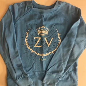 Blå sweatshirt med ZV-tryck - Blå sweatshirt med stort vitt ZV-tryck och krona framtill. Tröjan har rund halsringning, långa ärmar och ribbade muddar. Snygg detalj med glittrande prickar runt trycket.BARN STORLEK ÅR 10 ÅR