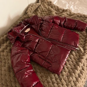 Vinröd pufferjacka från Nelly - Säljer en glansig vinröd pufferjacka från Nelly med huva och dragkedja framtill. Jackan har en oversized look, stora fickor och quiltad design. Perfekt för kalla dagar när du vill vara både varm och snygg. Den har ett litet hål i ena fickan men verkligen inget som man tänker på!❤️