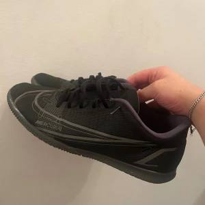 Nike Mercurial svarta fotbollsskor - Säljer ett par svarta Nike Mercurial fotbollsskor med grå detaljer och logga. Skorna har snörning, platt sula och rund tå. Ovandelen är i syntetmaterial och insidan är fodrad i ljuslila. Perfekta för inomhusspel och snabba vändningar.