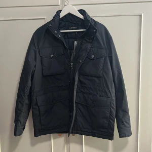 Mörkblå vadderad jacka J.Lindeberg storlek M - Snygg mörkblå vadderad jacka från J.Lindeberg med flera fickor framtill, hög krage och dragkedja. Perfekt för höst och vinter när du vill hålla stilen och värmen. Sprillans ny med prislapp och allting kvar. Orginalpris: 4700 kr