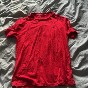 Röd t-shirt från Polo Ralph Lauren - Snygg röd t-shirt från Polo Ralph Lauren med klassisk rund hals och korta ärmar. T-shirten har en liten blå Polo-logga broderad på bröstet och är gjord i mjuk bomull som känns skön mot huden. Perfekt för dig som gillar stilrena och tidlösa plagg.