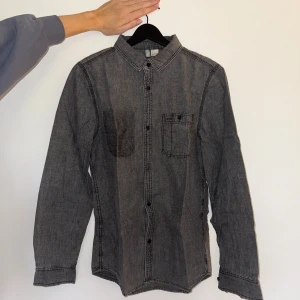 Grå skjorta från Divided H&M, stl S - Säljer en grå skjorta från Divided by H&M i storlek S. Skjortan har klassisk krage, svarta knappar och en bröstficka. Tillverkad i bomull och har långa ärmar med knapp vid manschetten. Perfekt till en avslappnad stil.