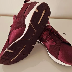 Vinröda sneakers från Puma strl 42 - Snygga vinröda Puma sneakers med vit sula och klassisk logga på hälen och plösen. Skorna har snörning och är tillverkade i syntetmaterial med textil och mockadetaljer. Modern design med rund tå och platt sula, perfekt för dig som gillar sportig stil.