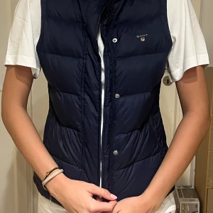 Mörkblå dunväst från GANT - Snygg mörkblå väst från GANT med quiltad design och ståkrage. Västen har dragkedja och tryckknappar framtill, samt två fickor på sidorna. Insidan är ljus med diskreta ränder i rosa och vitt. Perfekt för lager-på-lager och chill stil.