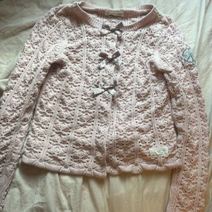 Rosa stickad kofta från Odd Molly - Superfin ljusrosa stickad kofta från Odd Molly med snyggt hålmönster och tre silkesrosetter framtill. Långärmad modell med ribbad kant och broderad logga på ärmen. Perfekt för dig som gillar söta detaljer och mjuka material.