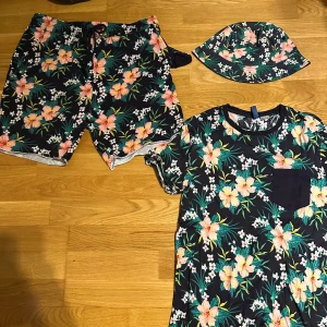 Blommig t-shirt från CHICCO, marinblå - Snygg marinblå t-shirt från CHICCO med tropiskt blommigt mönster i rosa, grönt och vitt. T-shirten har en rund hals, korta ärmar och en mörkblå bröstficka. Perfekt för sommarens chill dagar och strandhäng.