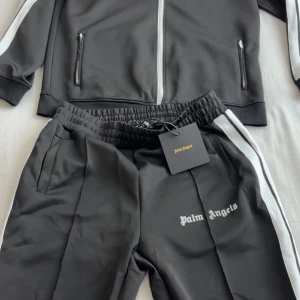 Palm Angels svarta mjukisbyxor med vit stripe - Svarta mjukisbyxor från Palm Angels med vit sidoremsa och logga tryckt på benet. Byxorna har elastisk midja med dragsko och raka ben. Tillverkade i polyester för en sportig vibe. Perfekta för en streetwear-look. Säljer då den ej passar mig 