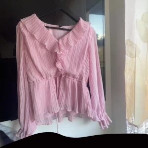 Ljusrosa blus med volang från Vero Moda - Säljer en ljusrosa blus från Vero Moda med plisserat tyg och volangdetaljer vid krage och framtill. Blusen har långärmade ärmar med resår vid handlederna och en feminin, flowy passform. Perfekt för dig som gillar romantiska vibes och mjuka färger.Vet inte vilken storlek de är men passar mig som har M