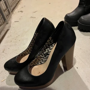 Svarta pumps med hög klack från VOX Shoes - Säljer ett par svarta pumps från VOX Shoes i storlek 36. Skorna har rund tå, hög träfärgad klack och ovandel i syntetmaterial. Insidan har ett grafiskt mönster i svart och vitt. Perfekta för dig som vill ha en klassisk men ändå cool look.