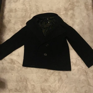 Svart skepparkavaj från Design by Si XS/S - Stilren svart skepparkavaj från Design by Si i storlek XS/S. Jackan har bred krage, dubbla knappar framtill och dekorativa knappar vid ärmslut. Tillverkad i 100% polyester med slät insida. Perfekt för dig som gillar klassisk och clean look.