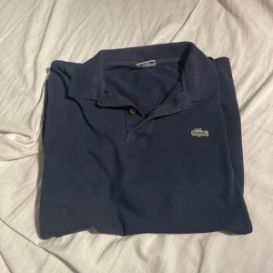 Mörkblå pikétröja från Lacoste - Säljer en klassisk mörkblå pikétröja från Lacoste med den ikoniska krokodilloggan på bröstet. Tröjan har korta ärmar, krage och knappar framtill. Perfekt för dig som gillar stilren och tidlös design. St S/M