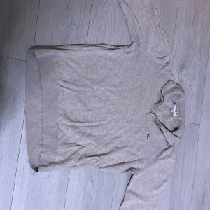Beige stickad half zip-tröja Lacoste - Säljer en beige stickad tröja från Lacoste med half zip och slim fit-passform. Tröjan har lång ärm, diskret rutmönster och klassisk krokodillogga på bröstet. Perfekt för dig som gillar stilren och sportig vibe.