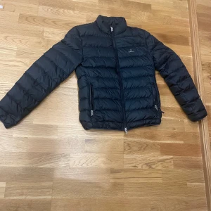 Svart pufferjacka från GANT XS - Säljer en svart pufferjacka från GANT i storlek XS. Jackan har horisontella sömmar, två dragkedjefickor och hög krage. Klassisk och stilren design med GANT-logga på bröstet. Perfekt för kyliga dagar.