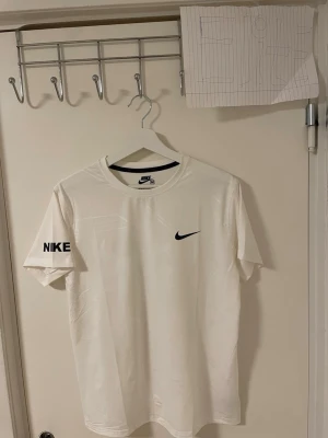 Vit Nike t-shirt   - Säljer en fräsch vit t-shirt från Nike i storlek M. T-shirten har korta ärmar och en klassisk rund hals. På bröstet finns en svart Nike Swoosh och på ena ärmen står det NIKE i svart. Materialet är mjuk bomull som känns skön mot huden.Liknande modell som originala.