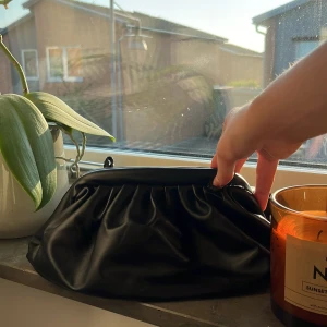 Svart kuvertväska i skinnimitation - Stilren svart kuvertväska med mjuk, rynkad design och klassisk metallstängning. Väskan är gjord i skinnimitation och har ett rymligt fack samt en innerficka med dragkedja för småsaker. Perfekt för dig som gillar minimalistisk och trendig look. Väskan är lite sliten och har ett hål på insidan och att öppningens mental grej har gått sönder men inget som syns på utsidan