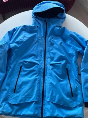 Blå skidjacka från Everest med huva strl L - Säljer en blå skid- och vindjacka från Everest i storlek L. Jackan är vind- och vattentät och har huva som funkar med hjälm, svarta dragkedjor och flera praktiska fickor framtill. Snölås, ventilation och snömuddar i ärmsluten gör den perfekt för skidåkning, blåsiga dagar och andra outdoor-aktiviteter.  Använd sparsamt under två säsonger. Nytvättad enligt instruktion.Inga skador, några ytliga smutsfläckar vid ärmslut och nedtill framtill (se bilder), inget som påverkar funktion eller helheten. 