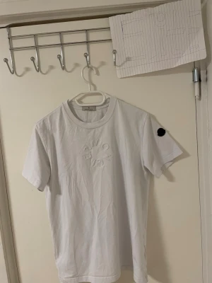 Vit Moncler t-shirt med logga - Säljer en clean vit t-shirt från Moncler med upphöjd logga på bröstet och svart patch på ärmen. Klassisk rund hals och korta ärmar. Materialet är mjuk bomull som känns skön mot huden. Perfekt för dig som gillar stilrena designer och exklusiva märken.Liknande modell som originala.
