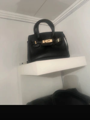 Svart handväska med gulddetaljer - Snygg svart handväska i skinn med klassisk form och guldfärgade metalldetaljer framtill. Väskan har två handtag och en elegant design som passar till många olika stilar. Perfekt för dig som gillar stilrena accessoarer med lyxig känsla. Aldrig använd, kom priv för mer bilder 