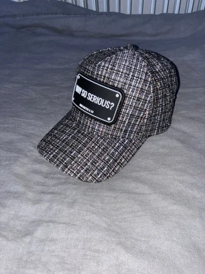 Rutig keps från John Hatter & Co - Säljer en snygg rutig keps från John Hatter & Co med texten 'WHY SO SERIOUS?' på en svart patch framtill. Kepsen har justerbar snapback och är i en mix av svart, grå och beige toner. Perfekt för att ge din outfit lite edge.