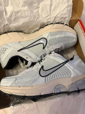 Nike Zoom Vomero 5 Pure Platinum STORLEK 38 - Nike Zoom Vomero 5 sneakers i färgen Pure Platinum/Chrome. Skorna har en mesh- och syntetöverdel med silvriga detaljer och svart Nike-swoosh. Platt sula med Cushlon-dämpning för extra komfort. Perfekt för dig som gillar stilrena och sportiga sneakers.