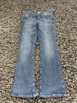 Blå bootcut jeans från Gina Tricot - Bootcut jeans i ljusblå tvätt från Gina Tricot, storlek 164. Jeansen har klassisk femficksdesign och snygga broderade detaljer med blommönster på bakfickorna. Tillverkade i jeansmaterial med knapp och dragkedja framtill.