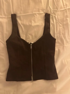 Svart korsett-topp från Zara - Säljer en svart korsett-topp från Zara med breda axelband och en snygg silverfärgad dragkedja framtill. Toppen har en figurnära passform och är tillverkad i ett mjukt syntetmaterial. Perfekt att styla med jeans eller kjol för en cool look.