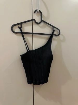 Svart ribbad croptop - Säljer en svart ribbad topp med snedskurning och smala axelband. Toppen är croppad och har en tight passform som framhäver siluetten. Perfekt för dig som gillar minimalistisk stil och vill ha något enkelt men ändå snyggt.