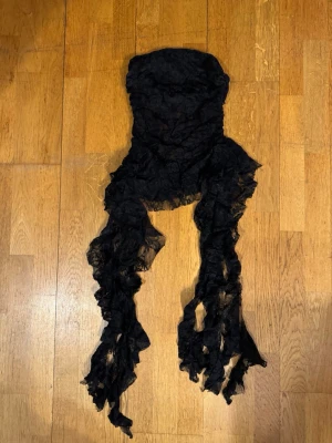Svart spetsväst med fransar från Emmabellin xs - Unik svart väst från Emmabellin i transparent spets med långa, dramatiska fransar framtill. Västen är strapless och har en cool, edgy look som sticker ut. Perfekt för dig som vill ha något extra till din outfit.