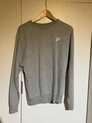 Grå sweatshirt från Nike, storlek S - Grå sweatshirt från Nike i storlek S. Tröjan har ett broderat Nike-logo på bröstet och är tillverkad i mjuk bomullsmix med ribbade muddar vid ärmslut och nederkant. Perfekt för chill eller träning, enkel och stilren design med rund hals.