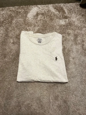 Ljusgrå t-shirt från Polo Ralph Lauren - Säljer en ljusgrå t-shirt från Polo Ralph Lauren i bomull med klassisk rund hals och svart broderad logga på bröstet. Sparsamt använd! Lite schysstare sandig färg på t-shirten otroligt sällsynt. Perfekt för dig som gillar stilrena och enkla plagg med snygg passform. Priset ej huggget i sten hör av er vid frågor eller funderingar! 
