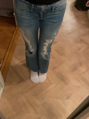 Blå ripped jeans från Gina Tricot Young - Säljer ett par blå jeans från Gina Tricot Young med slitna detaljer och hål på benen. Modellen har normal passform och bootcut 