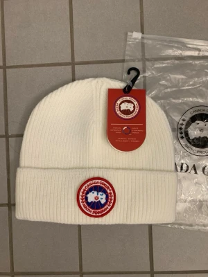 Vit Mössa - Säljer en vit mössa med klassisk rund patch framtill i rött, blått och vitt. Mössan är tillverkad i mjukt material och har en bred uppvikt kant. Perfekt för kalla dagar och snygg till vinterlooken.