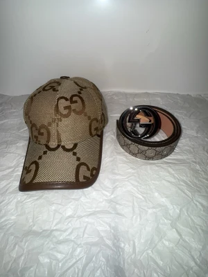 Beige GG-mönster keps och beige bälte  - Säljer en beige keps och bälte med klassiskt GG-mönster i brunt. Kepsen har bruna detaljer i skinn och justerbart band baktill. Materialet är bomull och skinn, och märket syns tydligt på insidan. Perfekt för dig som gillar lyxiga accessoarer.