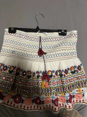 Broderad vit kortkjol med tofsar - Säljer en vit kortkjol med färgglada broderier och blommönster i rött, blått, gult och grönt. Kjolen har smock upptill, dekorativa tofsar och volangkant. Materialet känns som bomull och kjolen har en bohemisk vibe med detaljerade sömmar och spegelbroderier.  Snören sticker ut och har släppt 
