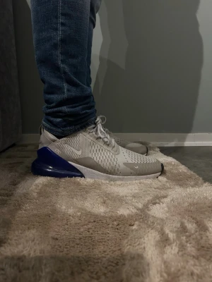 Nike Air Max 270 sneakers grå/blå - Nike Air Max 270 sneakers i ljusgrå mesh med vita detaljer och en stor blå Air-sula. Skorna har snörning, rund tå och platt sula. Snygg sportig design med ventilerande material och tydlig Nike-logga på sidan och hälen.