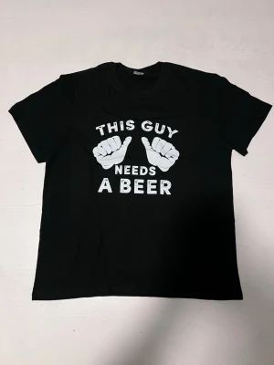 ”This guy needs a beer” T-shirt - En svart t-shirt med ett roligt och avslappnat budskap. Trycket visar två tummar som pekar mot texten “This guy needs a beer” i tydliga, vita bokstäver. Perfekt för vardag, fest eller som humoristisk present. Tröjan är i nytt skick och bekväm att bära.