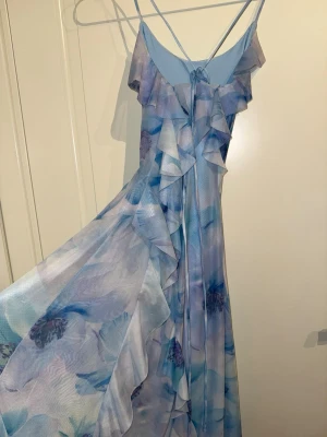Blommig blå långklänning med volanger - Säljer en superfin långklänning med blommigt mönster i olika blå nyanser och ljusa toner. Klänningen har tunna axelband, volangdetaljer längs fram och omlottkänsla. Materialet är lätt och luftigt, perfekt för varma dagar.