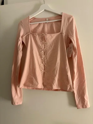 Ljusrosa långärmad topp från Nelly - Säljer en ljusrosa långärmad topp från Nelly i storlek M. Toppen har fyrkantig urringning och dekorativa rosetter framtill. Materialet är mjukt och lätt bomull, perfekt för dig som gillar söta detaljer och enkel stil.