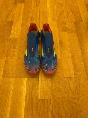 Adidas F50 blå fotbollsskor - Säljer ett par Adidas F50 fotbollsskor i blått med gula och rosa detaljer. Skorna har en strömlinjeformad design, platt sula med dobbar och klassiska Adidas-ränder på sidan. Insidan är röd och sulan har neonfärgade inslag. Perfekt för dig som vill sticka ut på planen. Skorna är använda ett få tal gånger och är riktigt snygga och i mycket bra skick.