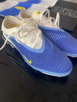 Blå och vita Nike fotbollsskor - Säljer ett par snygga Nike fotbollsskor i blått och vitt med gula detaljer. Skorna har snörning och är tillverkade i syntetmaterial med en platt sula och rund tå. Perfekta för dig som vill sticka ut på planen med stilrena färger och modern design.