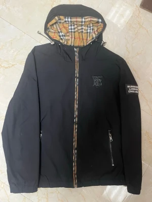 Svart Burberry vindjacka med huva - Svart vindjacka från Burberry med klassiskt rutigt foder i huvan och snygga logodetaljer på bröstet och ärmen. Jackan har dragkedja framtill, två fickor med dragkedja och elastiska muddar. Perfekt för dig som vill ha en stilren och ikonisk look.( obs att den har en liten defekt vid halsen på spännet bild kan skickas vid intresse)