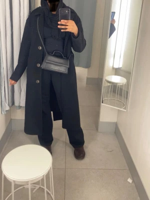 Svart lång kappa med knappar - Säljer en svart lång kappa med stora knappar framtill. Klassisk rak modell med långärmade ärmar och snygg krage. Perfekt för dig som gillar stilrena och tidlösa plagg. Kappan har en enkel och clean look som passar till många outfits.